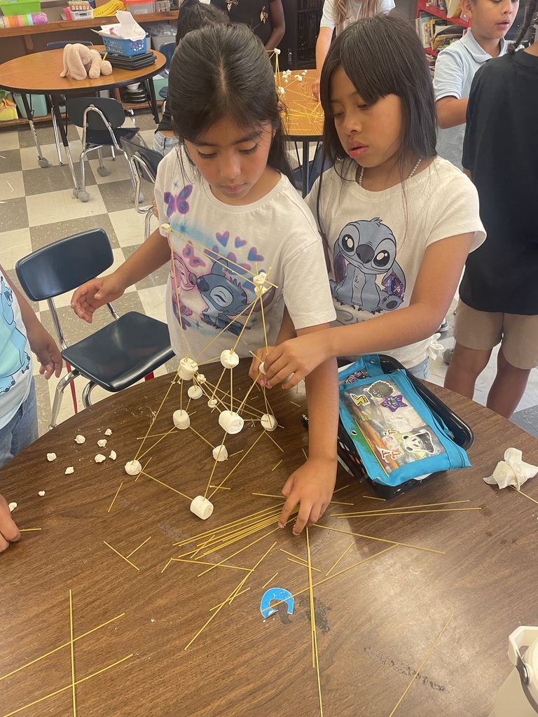 Two girls are standing a table with their tower made from spaghetti and marshmallows.