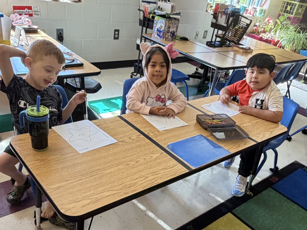 Three students are sitting at a table in their classroom coloring on paper.