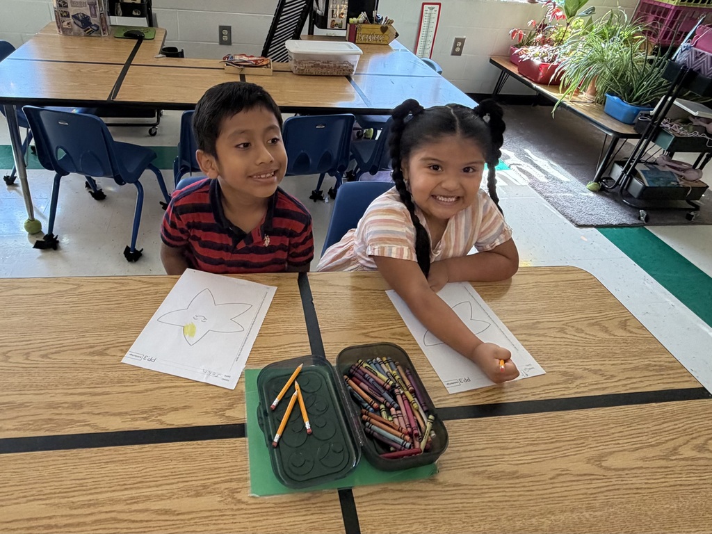 Two students are sitting at a table in their classroom coloring on paper.
