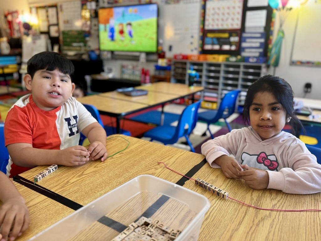Two students are sitting at a table in their classroom putting wooden letter beads onto string.