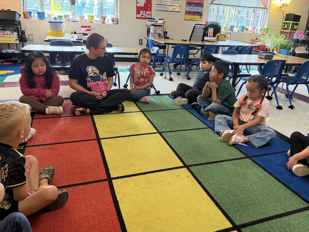 There are several students sitting around a carpet that has different color squares with an adult.