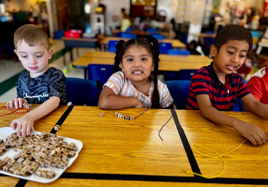 Three students are sitting at a table in their classroom putting wooden letter beads onto string.