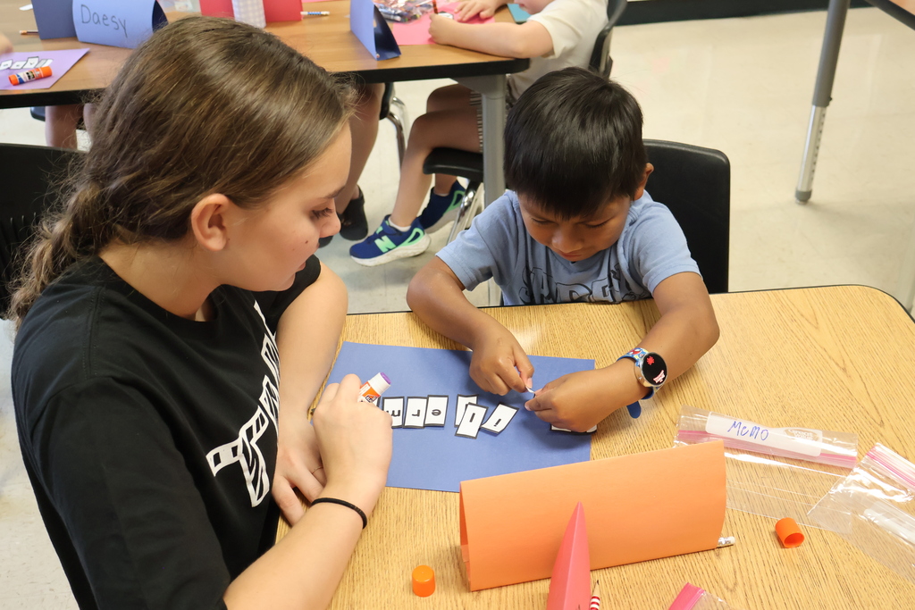 An assistant helps a student glue the letters of his name to a piece of construction paper. 