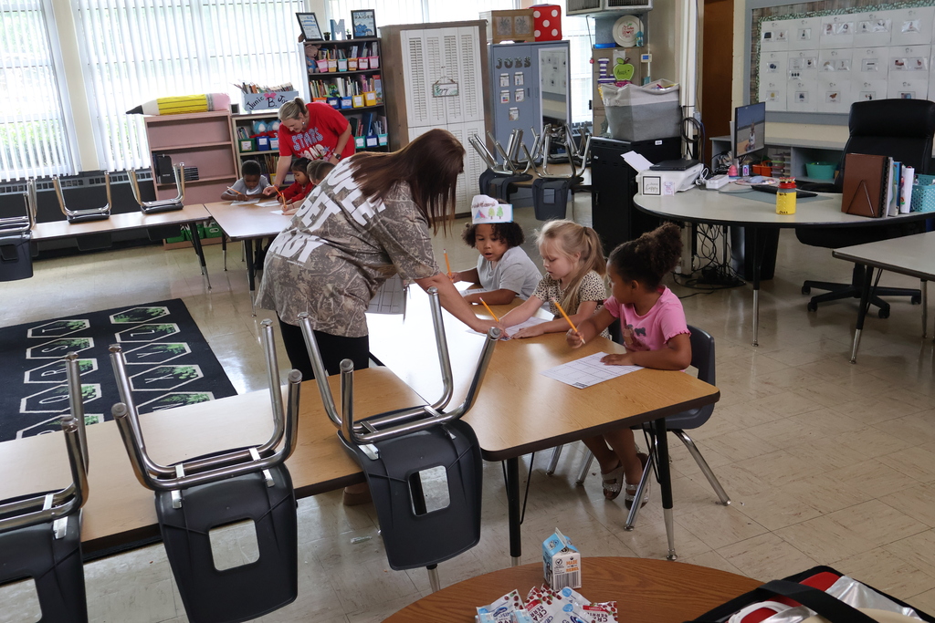 There are several students sitting down at two tables as two teachers walk around and help them with their work. 