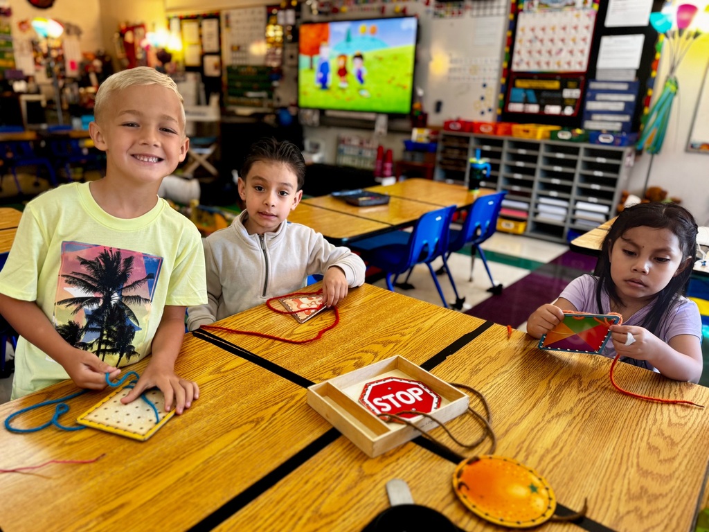 Three students are sitting at their desks as they put colored string through boards. 