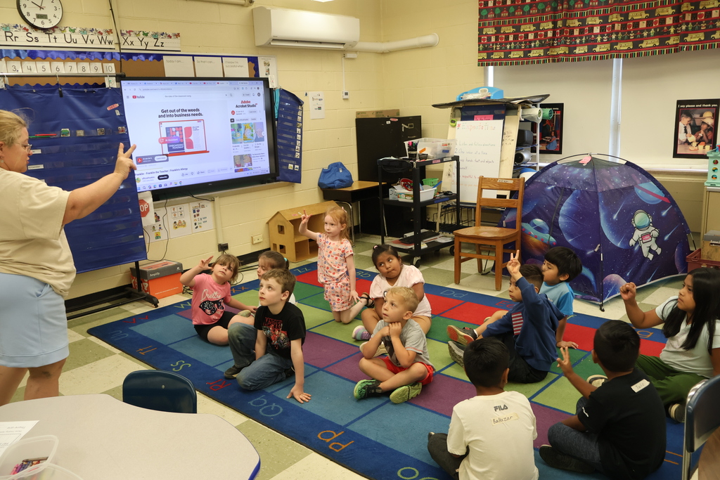 There are several students sitting on a colorful carpet while they each hold up two fingers like their teacher. 