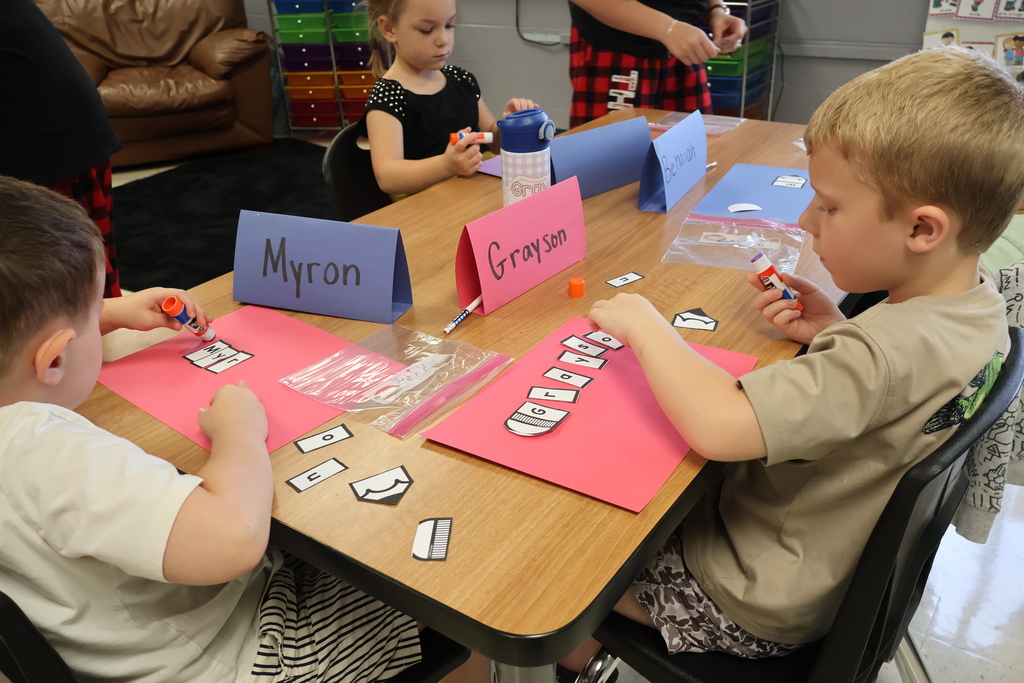 There are three students sitting at a table gluing pieces of paper to construction paper that spells their name according to their name tag in front of them. 