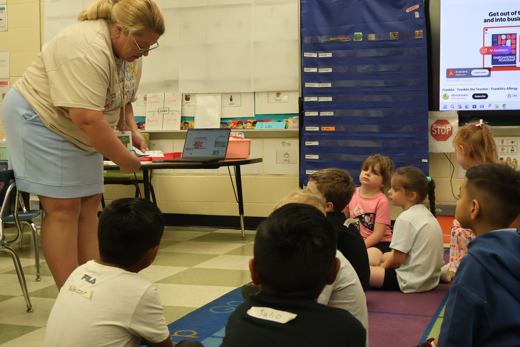 A teacher is demonstrating to students sitting on a colorful carpet how to cut paper. 