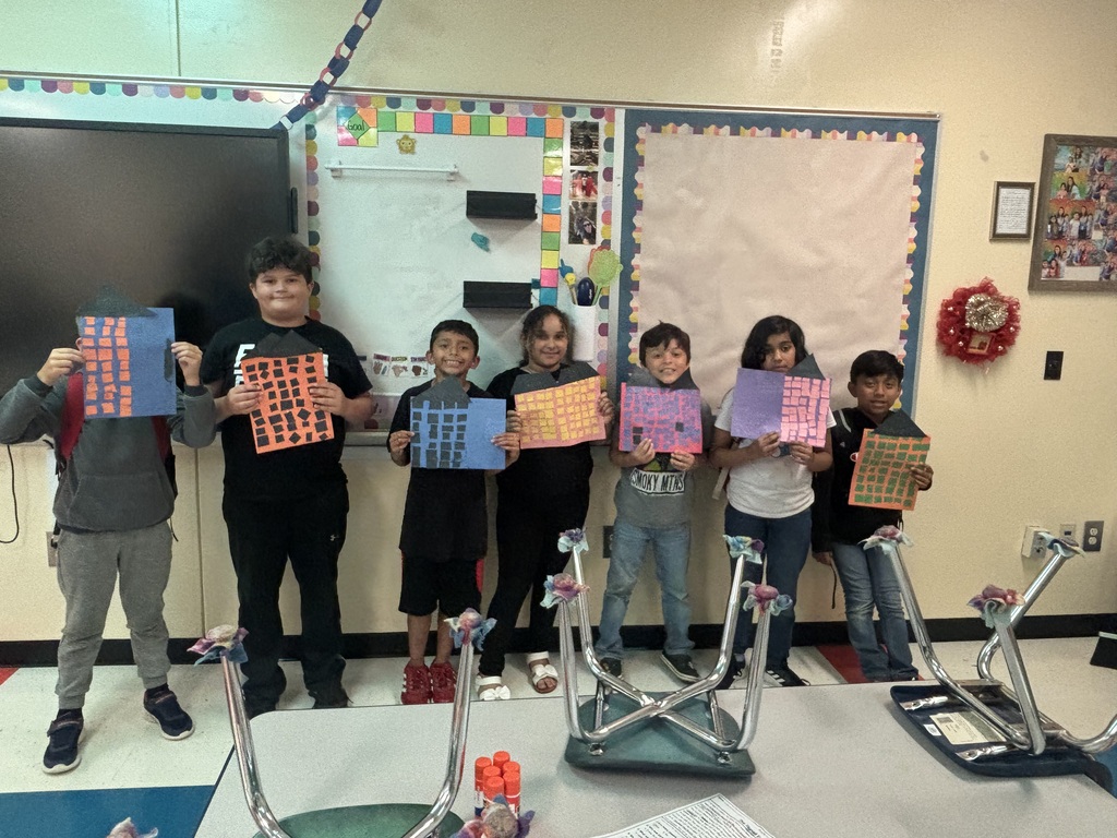 There are seven students standing in front of a white board holding up a piece of construction paper they used to create for their buildings. 