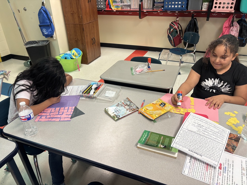 Students are sitting down at a table working on gluing pieces of square construction paper to another piece of construction paper. 