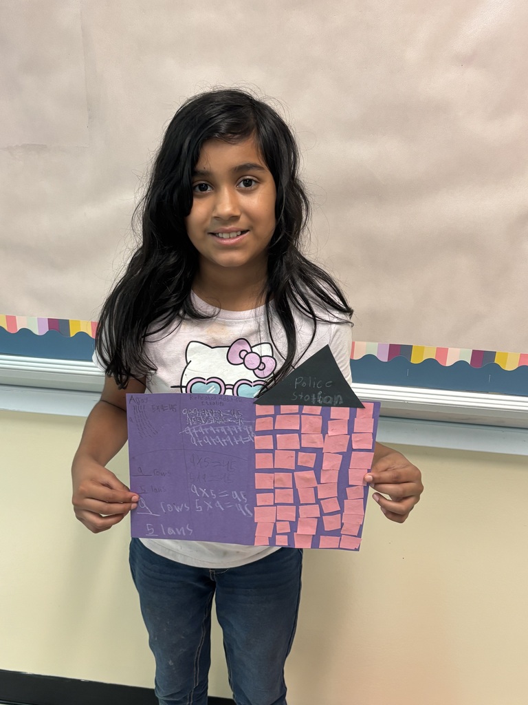 A student is standing in front of a bulletin board holding up a piece of construction paper they used to create for their buildings. 
