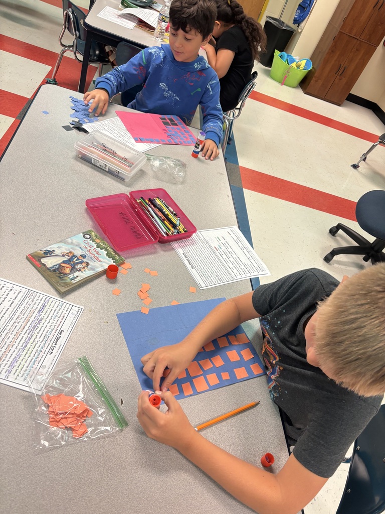 Students are sitting down at a table working on gluing pieces of square construction paper to another piece of construction paper. 