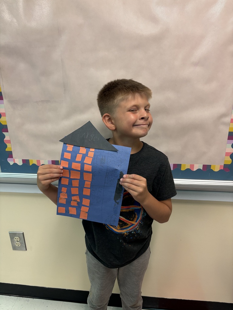 A student is standing in front of a bulletin board holding up a piece of construction paper they used to create for their buildings. 