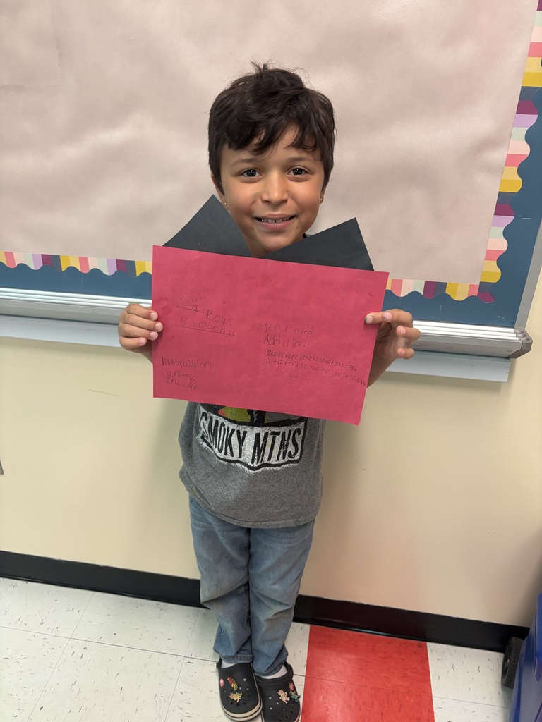 A student is standing in front of a bulletin board holding up a piece of construction paper they used to create for their buildings. 