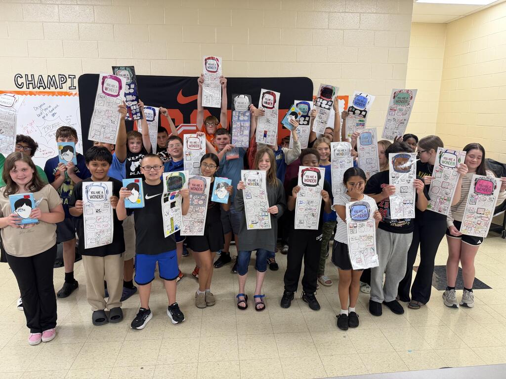 There is a group of students all posed together for a picture holding up a piece of paper with a robot on it they decorated. 
