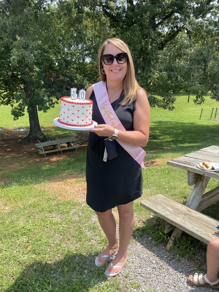 A teacher is standing outside holding a birthday cake with candles that spell out 50. 