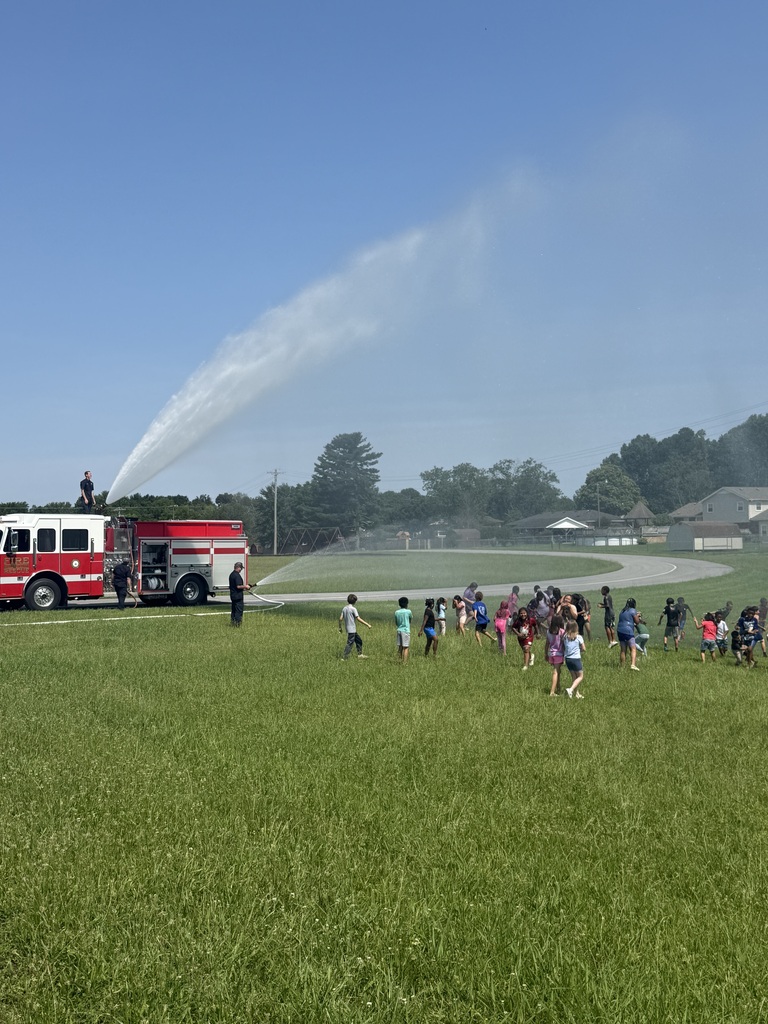 Students are outside playing in the grass with water that is being sprayed from a fire truck.