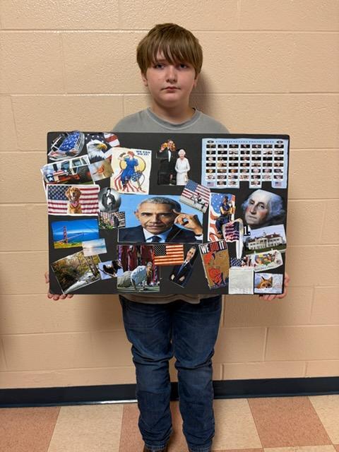A student is standing in the hallway holding up his American-themed collage poster. 