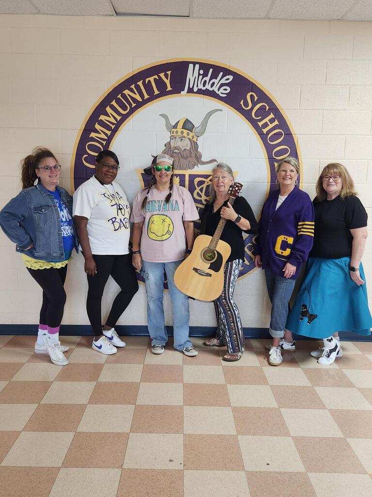 Six teachers are standing in the hallway posed for a picture wearing their outfits for "decades day". 
