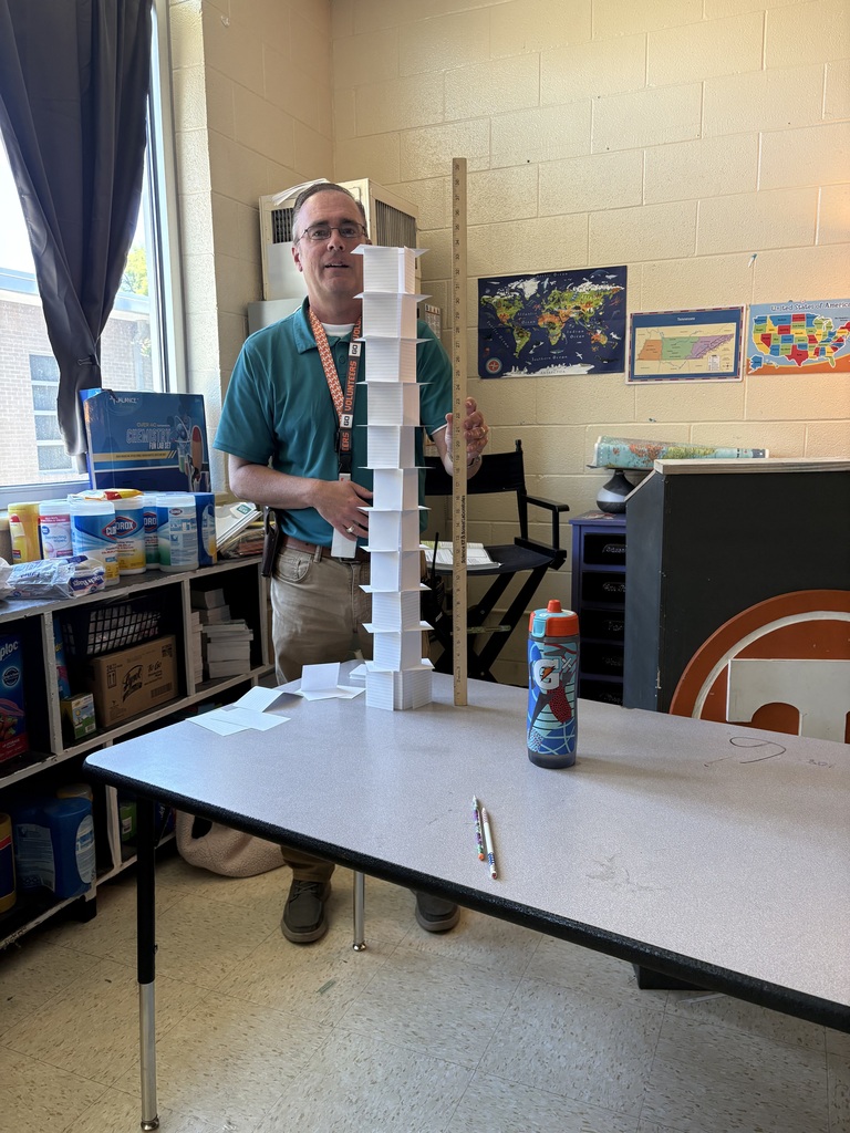 The teacher stands behind a table with a tower made of index cards holding a yard stick to measure it. 
