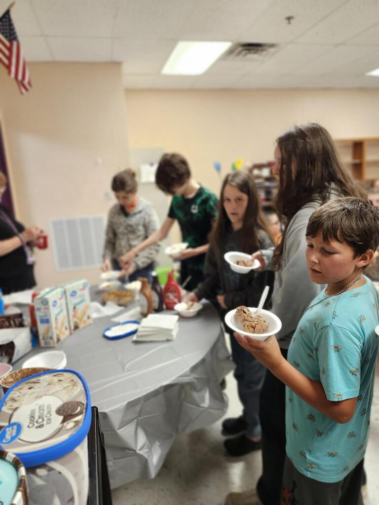 There are several students along with their teacher gathered around a table putting ice cream into a bowl. 