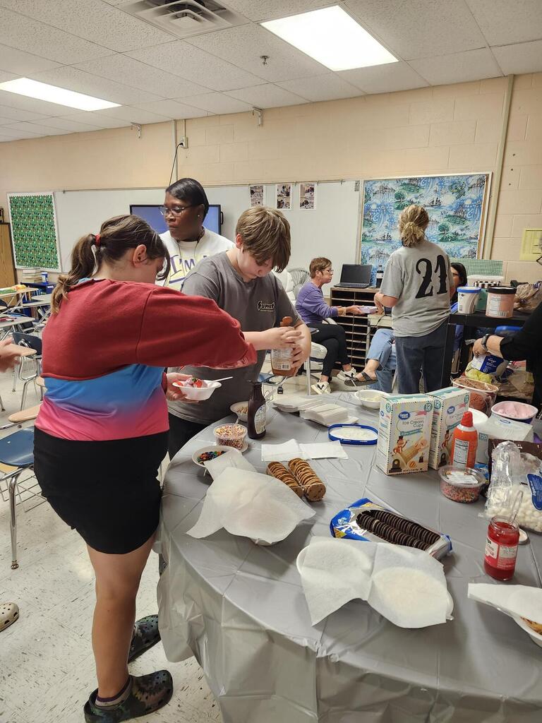 There are several students along with their teacher gathered around a table putting ice cream into a bowl. 