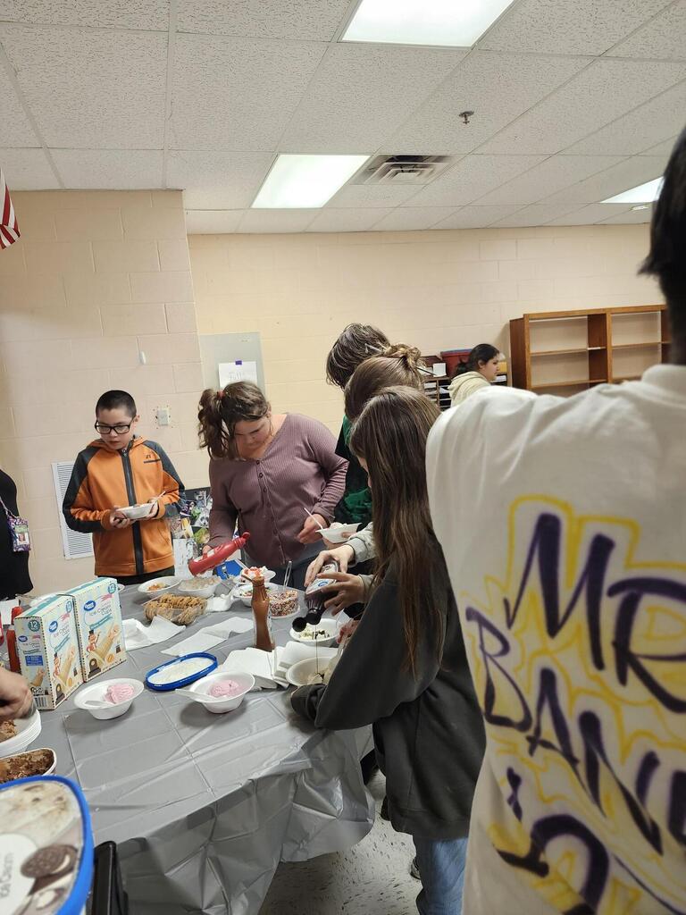 There are several students along with their teacher gathered around a table putting ice cream into a bowl. 