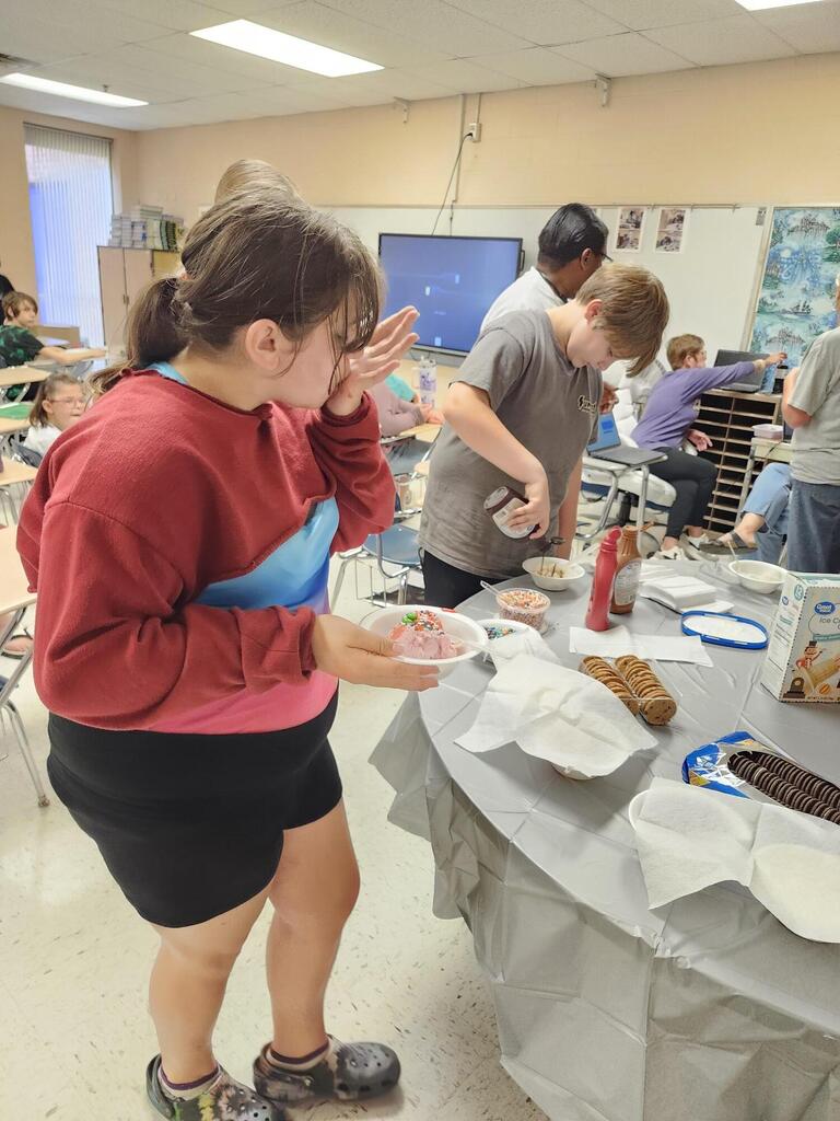 There are several students along with their teacher gathered around a table putting ice cream into a bowl. 