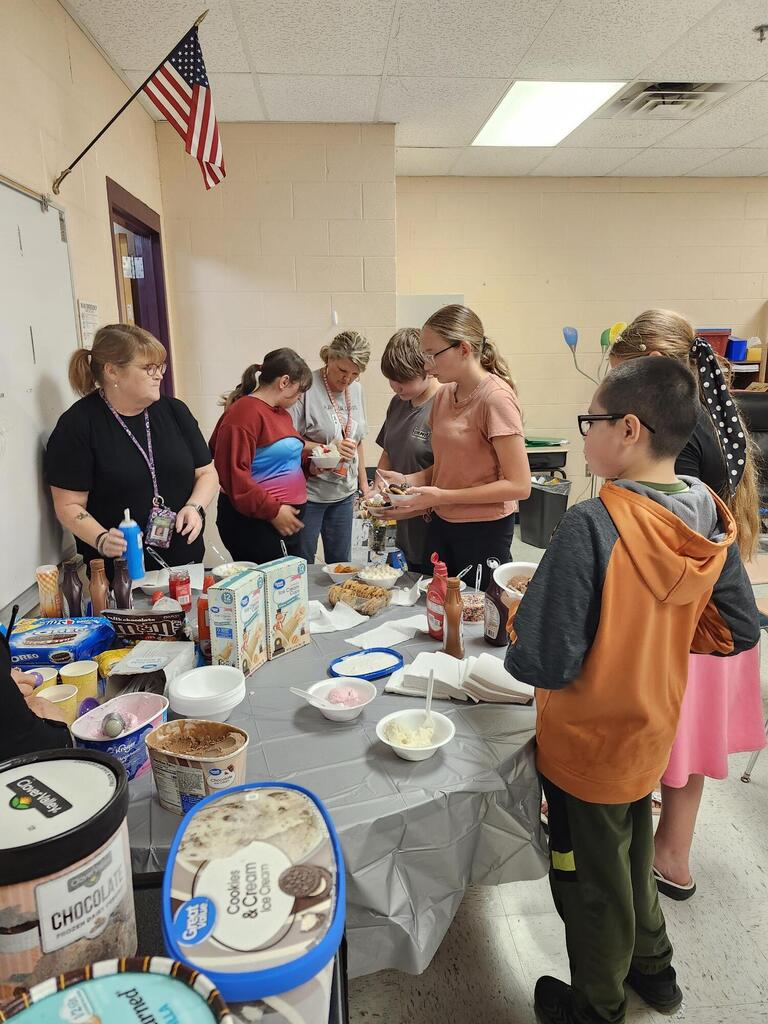 There are several students along with their teacher gathered around a table putting ice cream into a bowl. 