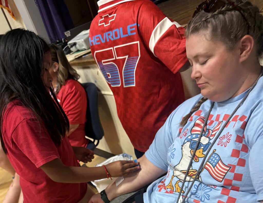 A teacher is sitting in a chair as a student puts a temporary tattoo on her arm. 