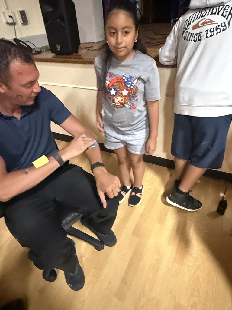 The school resource officer is sitting in a chair as a student puts a temporary tattoo on his arm. 