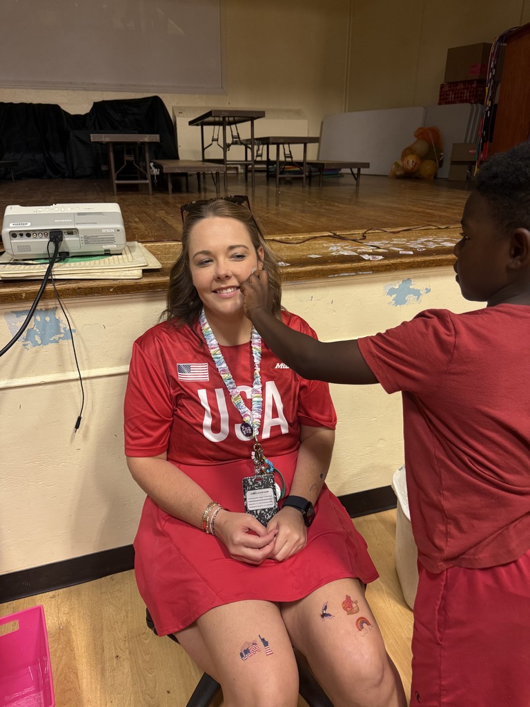 A teacher is sitting in a chair as a student puts a temporary tattoo on her face. 