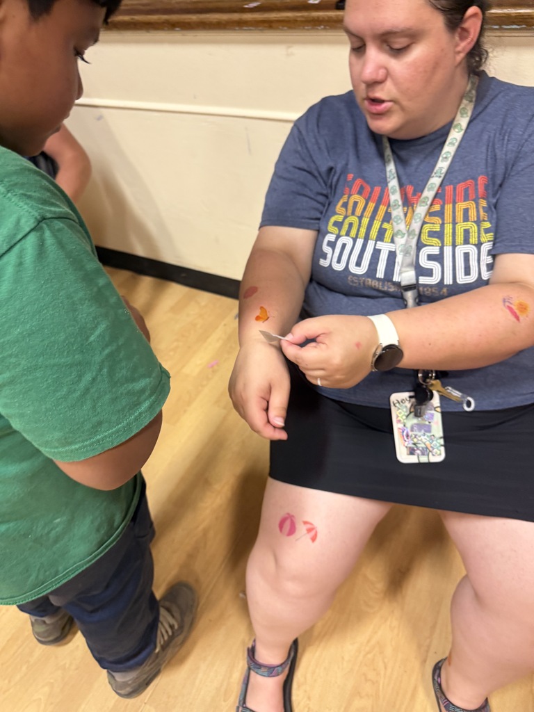 A teacher is sitting in a chair as a student puts a temporary tattoo on her arm. 