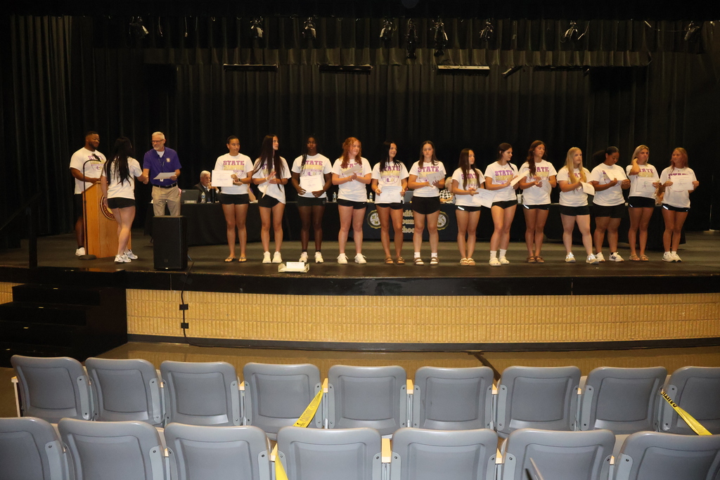 Players from the Community High School Viqueens softball team are called on stage by their coach to receive a certificate from Superintendent Tim Harwell.  The players are all standing in a line on stage in front of the school board members.
