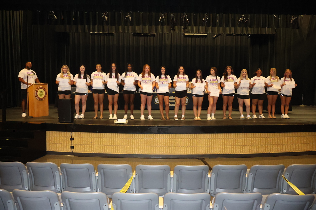 Players from the Community High School Viqueens softball team players are all standing in a line on stage in front of the school board members while holding their certificates. 