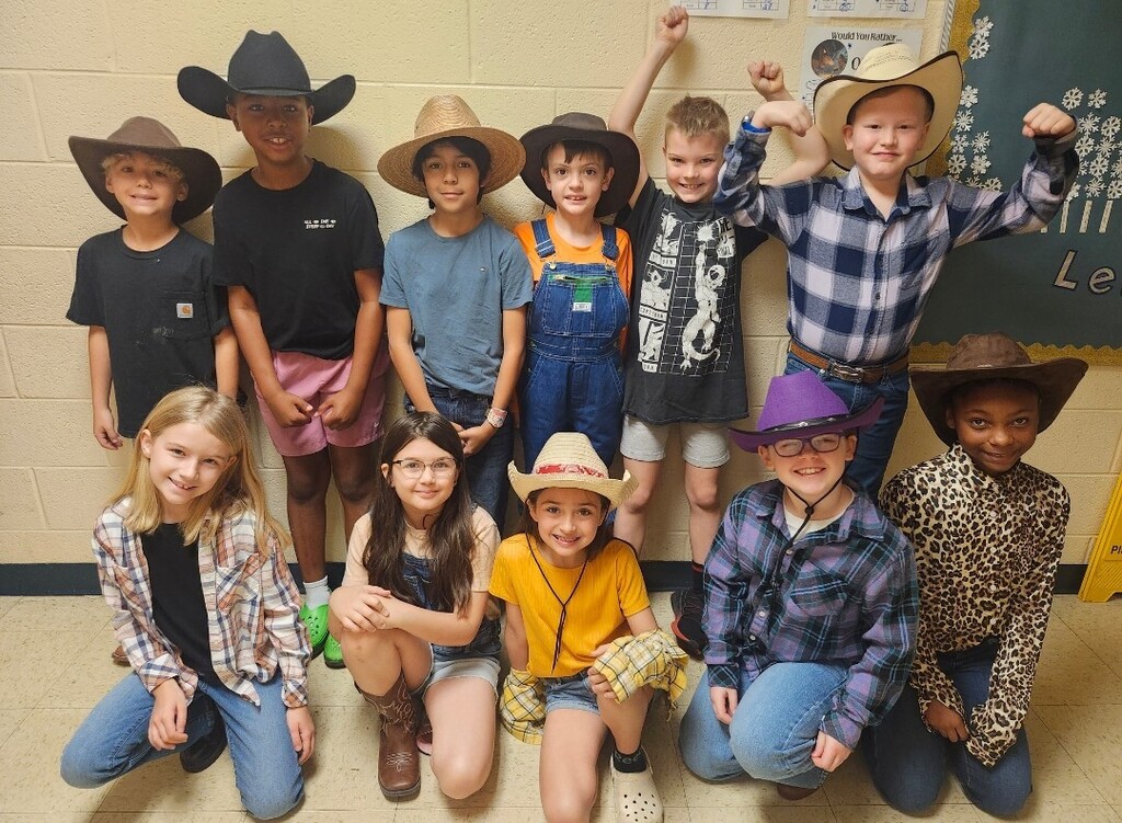 There is a group of students standing in the hallway posed for a picture wearing cowboy boots and hats.