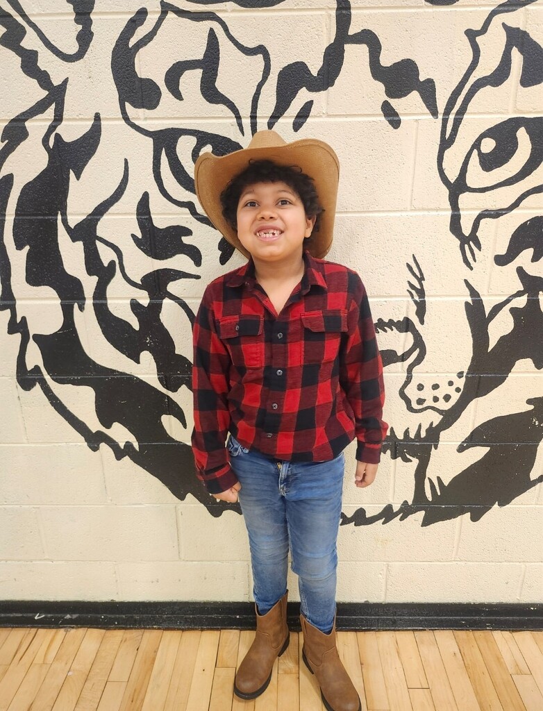 A student wearing cowboy boots and a hat is standing in the hallway for a picture in front of the school's tiger logo.