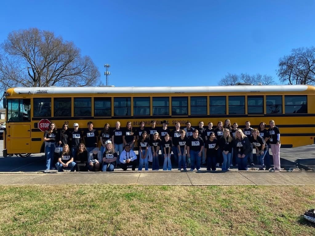 There is a large group of students all wearing the same black shirt posed for a group picture in front of a school bus. 