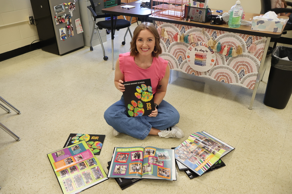 A teacher is sitting on the floor holding a yearbook in her hands with others in front of her on the floor open and fanned out. 