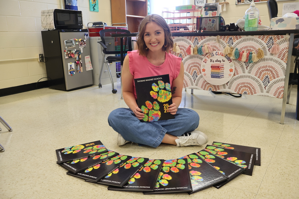 A teacher is sitting on the floor holding a yearbook in her hands with others in front of her on the floor fanned out. 