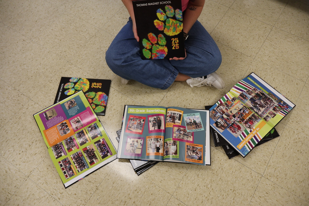 A teacher is sitting on the floor holding a yearbook in her hands with others in front of her on the floor open and fanned out. 