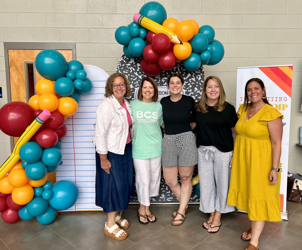 Five educators are standing for a group picture in front of a school-themed background that has items such as balloons. 