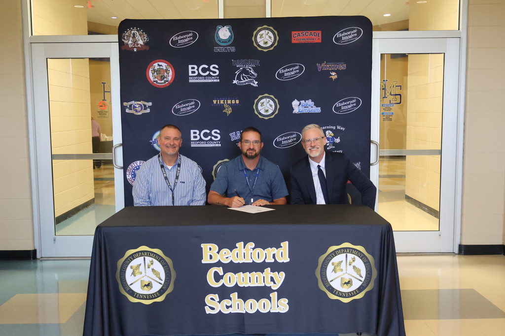 There are two people seated at a table covered in a black Bedford County Schools tablecloth all in front of a backdrop with all the schools logos on it. 