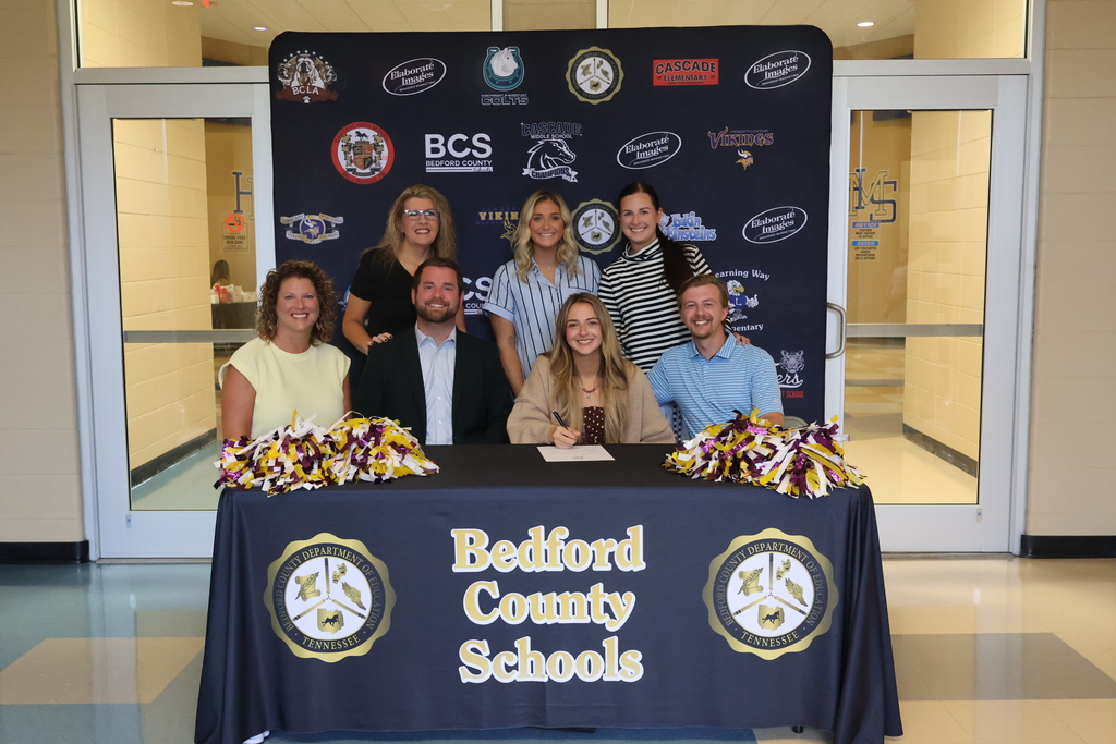 There are four people seated at a table covered in a black Bedford County Schools tablecloth along with three others standing behind them all in front of a backdrop with all the schools logos on it. 