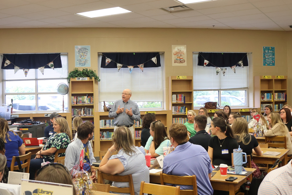A special speaker stands in front of a library full of people seated at tables as he speaks to them. 