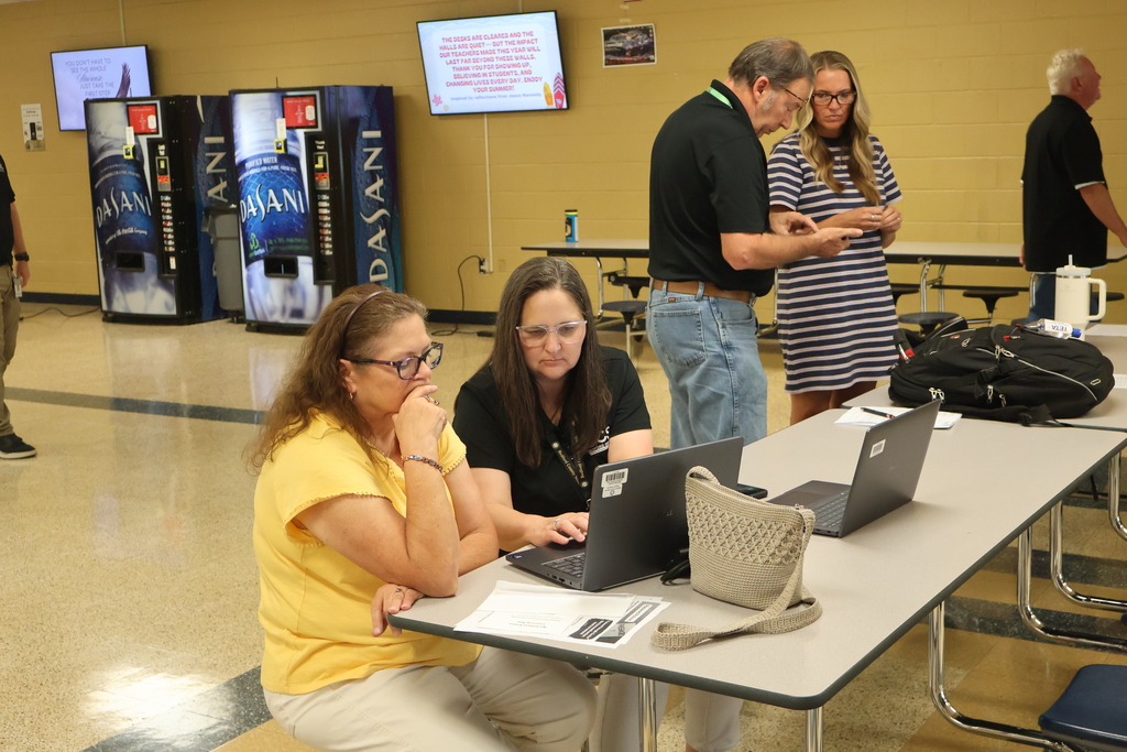 The Director of Technology is seated at a table helping a new teacher with her laptop while another person from that department also helps a new teacher. 