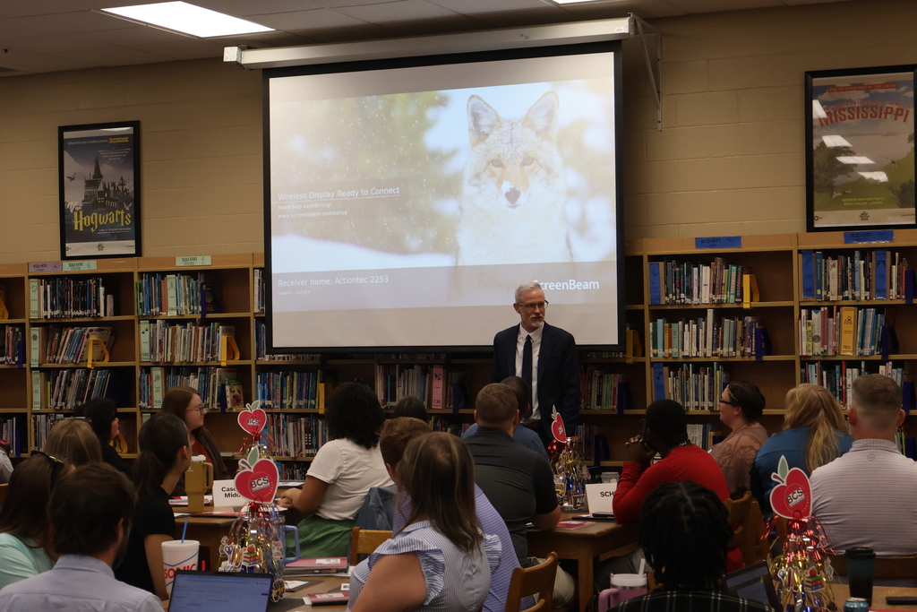 The Superintendent, Mr. Tim Harwell, stands in front of a library full of people seated at tables as he speaks to them. 