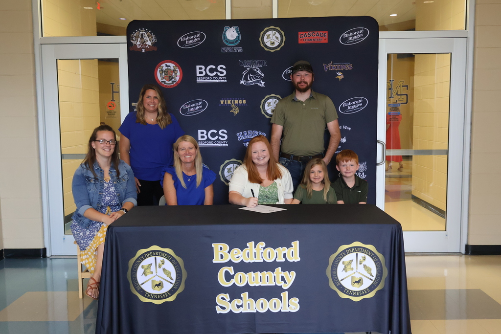 There are five people seated at a table covered in a black Bedford County Schools tablecloth along with two others standing behind them all in front of a backdrop with all the schools logos on it. 
