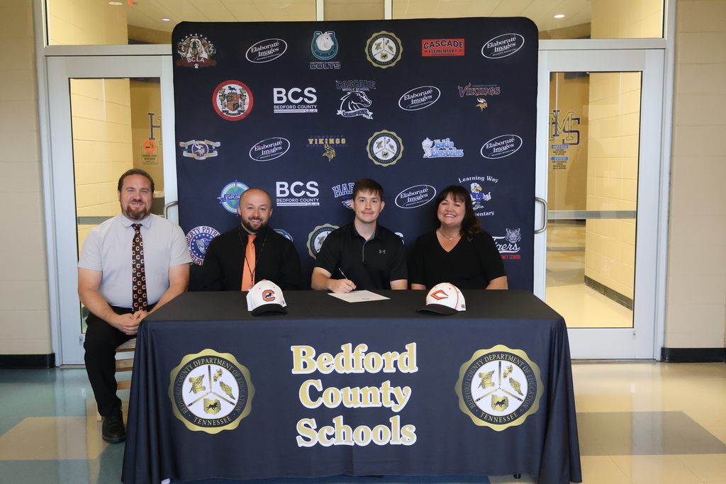 There are four people seated at a table covered in a black Bedford County Schools tablecloth all in front of a backdrop with all the schools logos on it. 