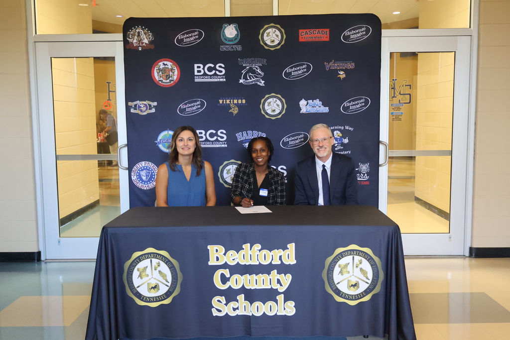 There are three people seated at a table covered in a black Bedford County Schools tablecloth all in front of a backdrop with all the schools logos on it. 